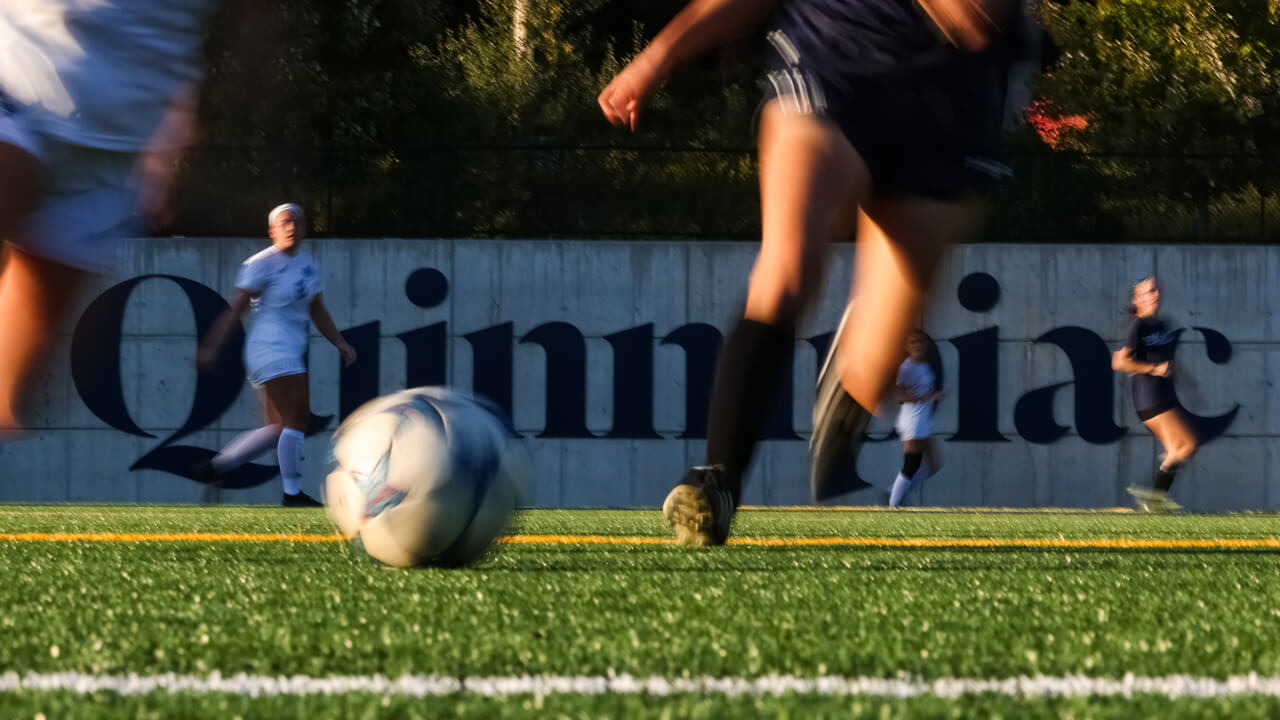 Soccer players run in a blur with a ball across the field with a Quinnipiac sign in the background