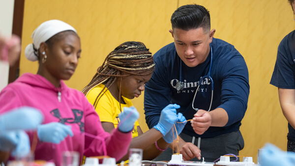 Two high school students wear surgical gloves and work with a faculty member on a health sciences skill