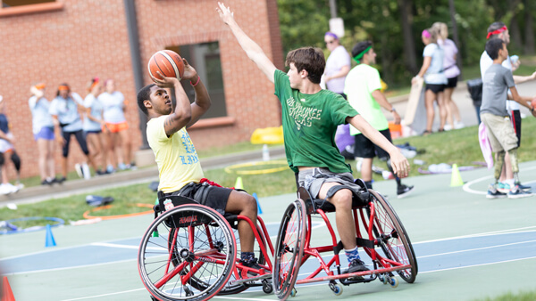 Two young people with limb loss in wheelchairs play basketball outside