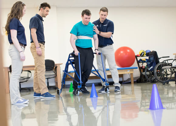 A person using a walker performs an exercise with the help of physical therapy students