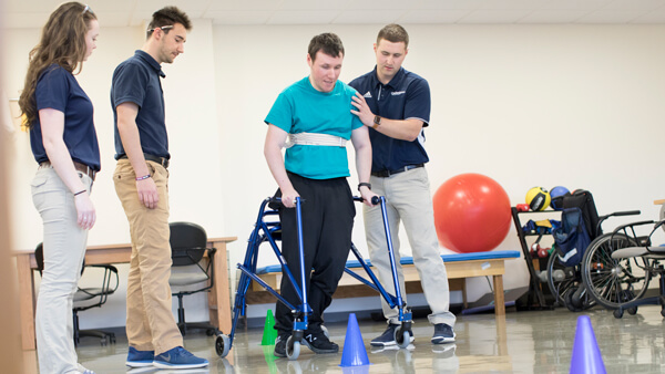 A person using a walker performs an exercise with the help of physical therapy students
