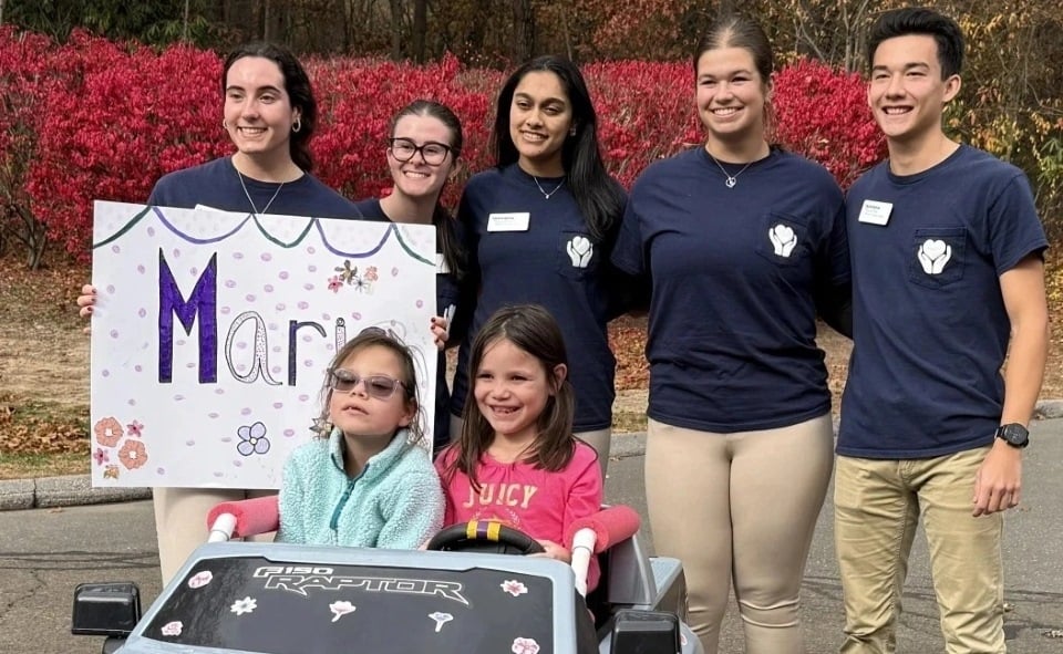 Physical therapy students pose behind two children in a Go Baby Go car.